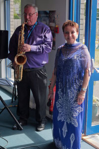 A woman in a long dress smiling at the camera next to a man playing the saxophone