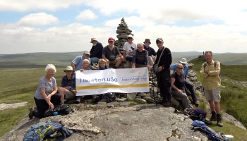 Eleven people stand at the top of a hill, with a view of fields behind them, holding a Tiverton u3a sign