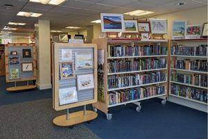 A library. To the left of a bookshelf is a stand with artwork on and paintings and drawings are on top of the bookshelves too.
