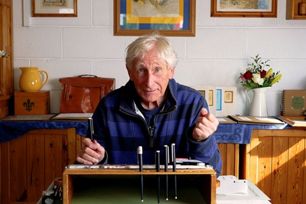 A man in a blue and black striped jumper is sitting at a desk in his home. The desk is slightly slanted and there are pens on it. The man is holding one pen above a piece of paper with ink on it, and is looking directly at the camera.