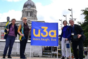 Four people stood in front of an old white building. They are holding a blue flag with 'u3a' 'learn, laugh, live' written on it in yellow writing.