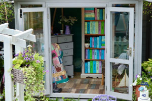 A shed with open doors reveals a set of drawers, on top of which are several items including a purple jug with flowers in it. Next to the drawers is a brightly coloured quilt and a pair of walking boots. There is a book case with brightly coloured model books, all of which with words on that represent an activity. There is a wicker chair and a model boat in the other corner of the shed. The shed is surrounded by leaves and flowers, including a hanging flower basket.
