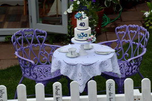 A table covered with a white table cloth which has on it two cups and saucers and in the centre of the table, a white two tiered cake. On the top tier, it says "40" and on the bottom tier "u3a Southport". The cake has flowers on the top and around the cake platter. There are two purple seats around the table.