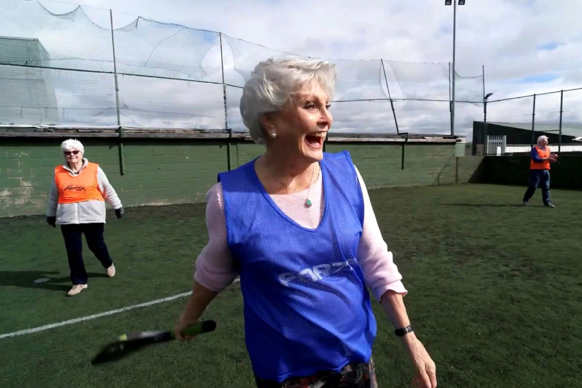A woman (Angela Rippon) wearing a blue sports bib, standing on a cricket pitch. She is holding a bat and is moving - she is smiling broadly. In the background, you can see another smiling woman in an orange bib.