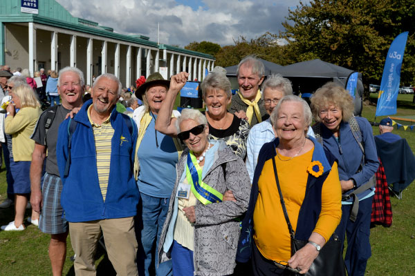 Nine people, all wearing shades of yellow and blue, are standing at an outside event. They are huddled together, facing the camera, and all are wearing big smiles. There are blue u3a flag banners in the background.