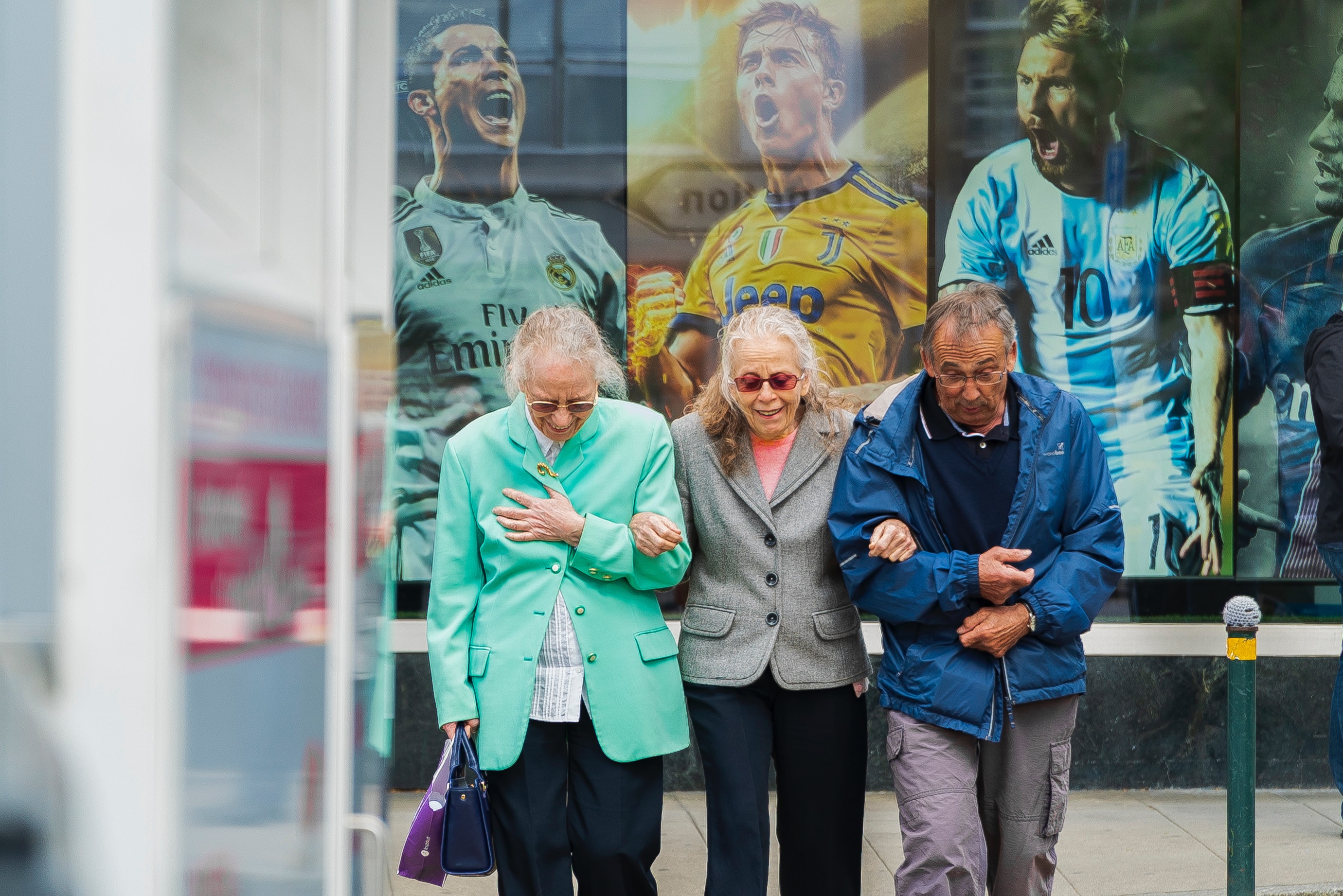 Three older people linking arms and smiling, walking away from a poster of images of three male footballers