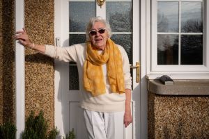 A woman wearing a white top, sunglasses and yellow scarf is standing in front of the white front door of a house