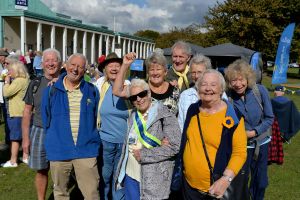 Nine people, all wearing shades of yellow and blue, are standing at an outside event. They are huddled together, facing the camera, and all are wearing big smiles. There are blue u3a flag banners in the background.