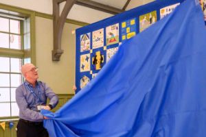 A man wearing a blue u3a lanyard pull a fabric covering off a quilt