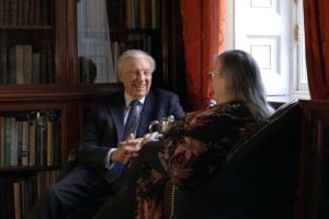 A man and woman are talking in a library. The man is facing the woman and is smiling. The woman's back is to us.