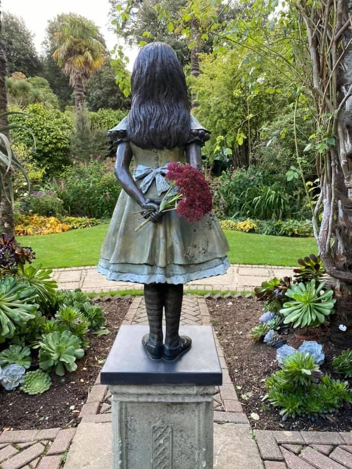 the back of a solid metal statue of a girl, with her hands behind her back. In the hands are red cut flowers. 