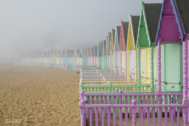 pastel pink green and yellow beach huts with sand on the left, in mist
