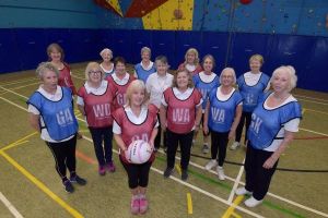 Women standing inside on a netball pitch; some are wearing blue bibs, some are wearing red bibs. The woman in the centre is holding a ball. They are all facing towards the camera and smiling.