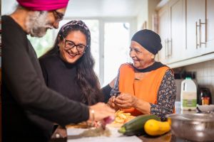 Two older adults with a younger relative cook in the kitchen. The older man is wearing a pink turban and is cutting vegetables. The older woman is peeling vegetables. They are smiling at each other.