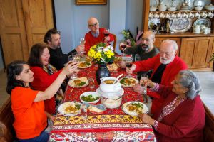 Seven people around a table which has a brightly coloured tablecloth on it. They each have a plate of curry - and in the middle is a white dish with a curry in it. They are holding up their glasses to each other.