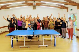 A large group of people standing in a hall, behind a table tennis table, with their hands outstretched in the air