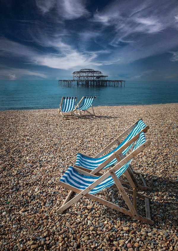 4 empty deckchairs on a pebble beach, with a dramatically dark sky and the skeleton of a burnt out pier in the distance 
