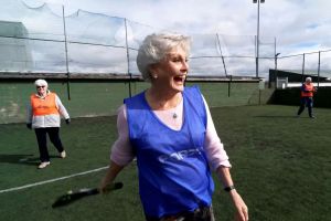 A woman (Angela Rippon) wearing a blue sports bib, standing on a cricket pitch. She is holding a bat and is moving - she is smiling broadly. In the background, you can see another smiling woman in an orange bib.